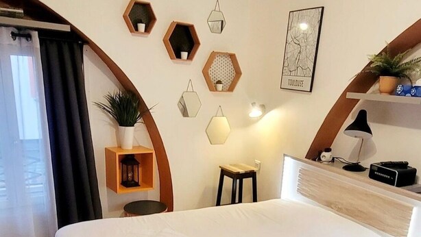 Une chambre d’hôtel avec un grand lit, un bureau et une petite table. Le sol est recouvert de carreaux hexagonaux, et les murs sont ornés d’étagères hexagonales sur lesquelles sont disposés divers objets.