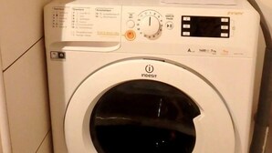 Une petite machine à laver, dotée d’un panneau de commande, d’un fer à repasser à vapeur et d’un rideau à rayures, dans un petit espace dédié à la buanderie.