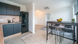 A modern kitchen with dark cabinets, a black refrigerator, and a wooden countertop. There is a dining area with a table and chairs, and a bowl of fruit on the table. - Carré d’As - Appartement de standing (Le Bourget)