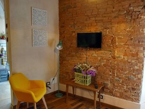 Ein Zimmer mit Backsteinwand, einem Flachbildfernseher über einem Holztisch mit Blumenvase, einem gelben Sessel und einer Stehlampe.