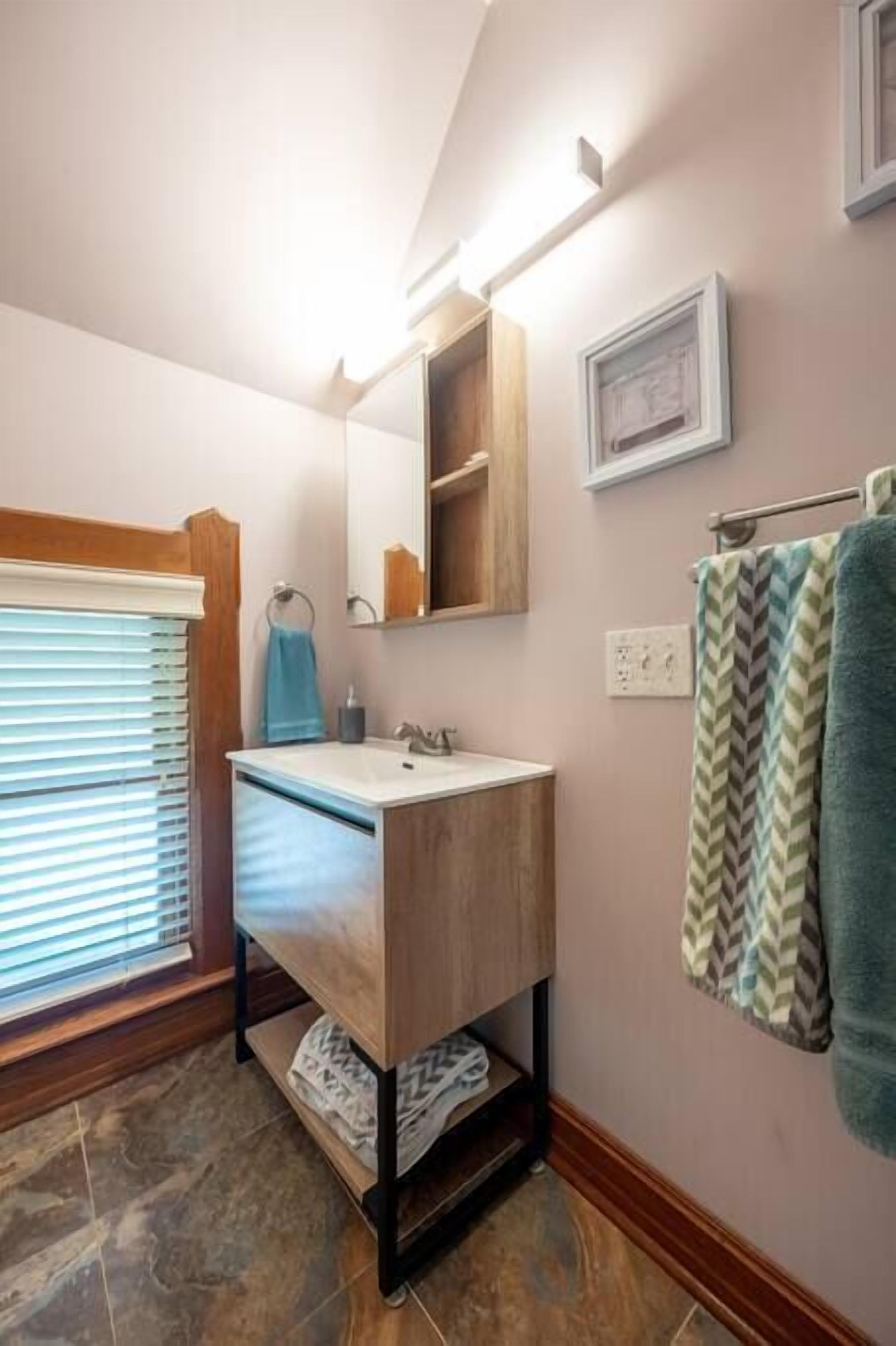 Un baño con tocador de madera, un espejo, toallero y una ventana con persianas.