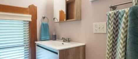 Un baño con tocador de madera, un espejo, toallero y una ventana con persianas.