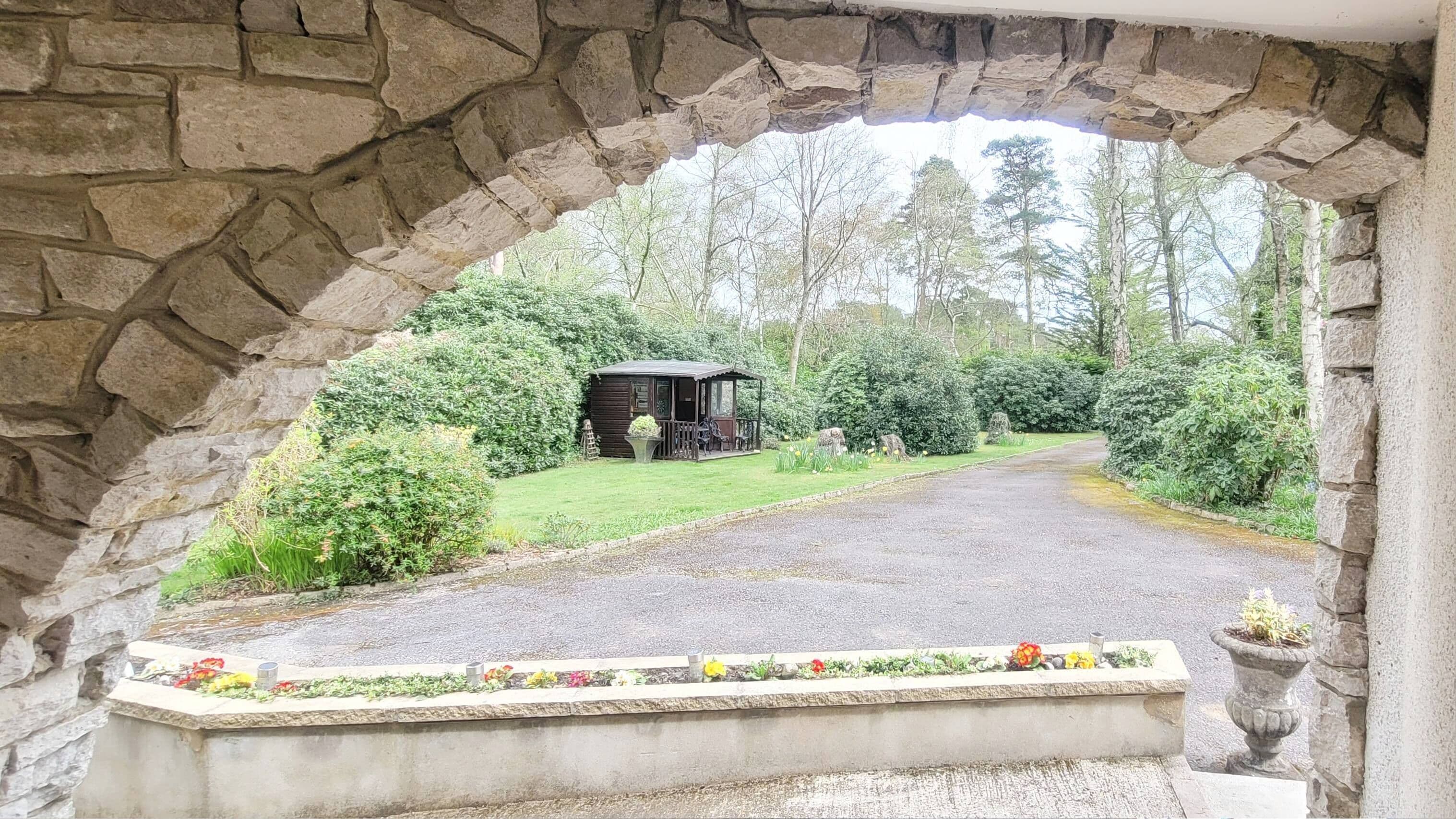 A stone archway leading to a small wooden structure surrounded by lush greenery.