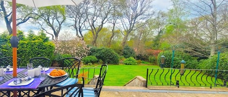 An outdoor patio with a table set for two, surrounded by chairs, and a view of a well-maintained garden with trees and shrubs.