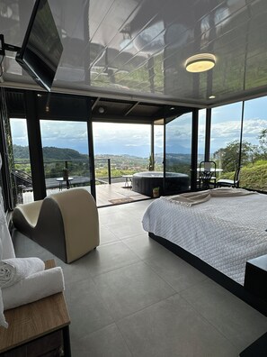 Een moderne slaapkamer met een groot bed, een bank, een klein tafeltje en een televisie. Er zijn grote ramen met uitzicht op de omgeving.