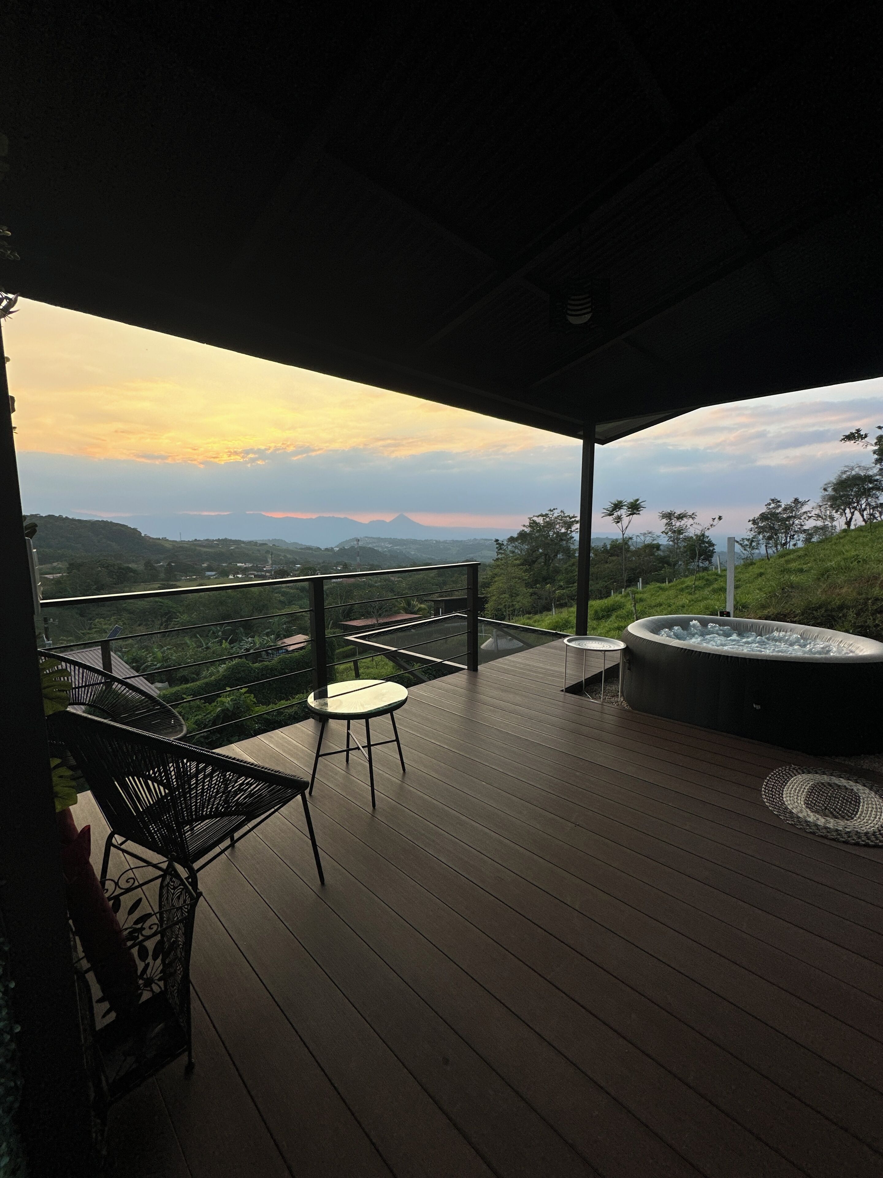 Una terraza de madera con un jacuzzi, una mesa y sillas, con vistas a un paisaje pintoresco con colinas y una montaña a lo lejos.