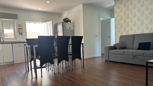 A modern kitchen with white cabinets and a built-in dishwasher, a dining area with black chairs, and a grey sofa in a living room with patterned wallpaper. - Moradia Privada COM Piscina Perto DA Praia (Apúlia)