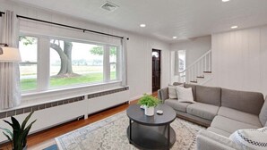 A modern living room with a large window, a grey sofa, a round coffee table, and a staircase in the background.