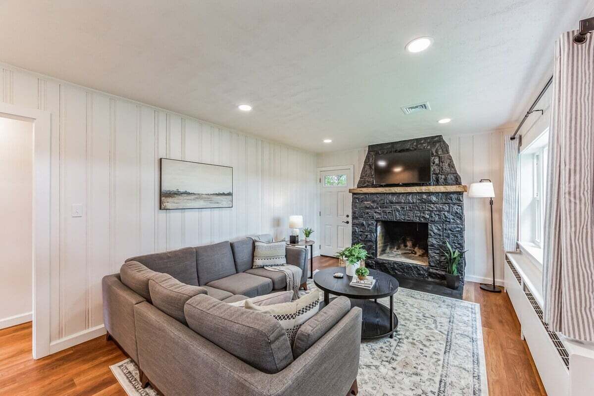 Een woonkamer met een stenen openhaard, een grijze bank, een ronde salontafel en een groot raam met gordijnen.