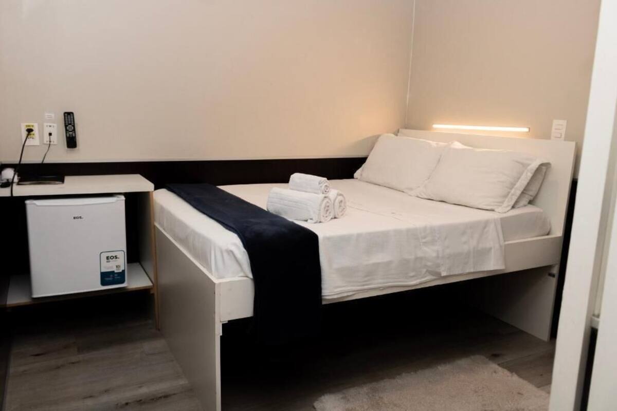 Habitación de hotel con una cama, una mesita de noche, un escritorio pequeño con una computadora y un panel de control en la pared.
