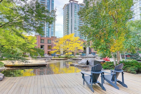 A wooden deck with two chairs overlooking a pond, surrounded by trees and buildings.