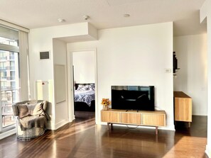 A modern living room with a flat-screen TV on a wooden TV stand, a comfortable armchair, and a view of a bedroom through an open door. - Entire Corner Unit in Prime Toronto Location. Steps from Subway, Shops & Dining (Toronto)