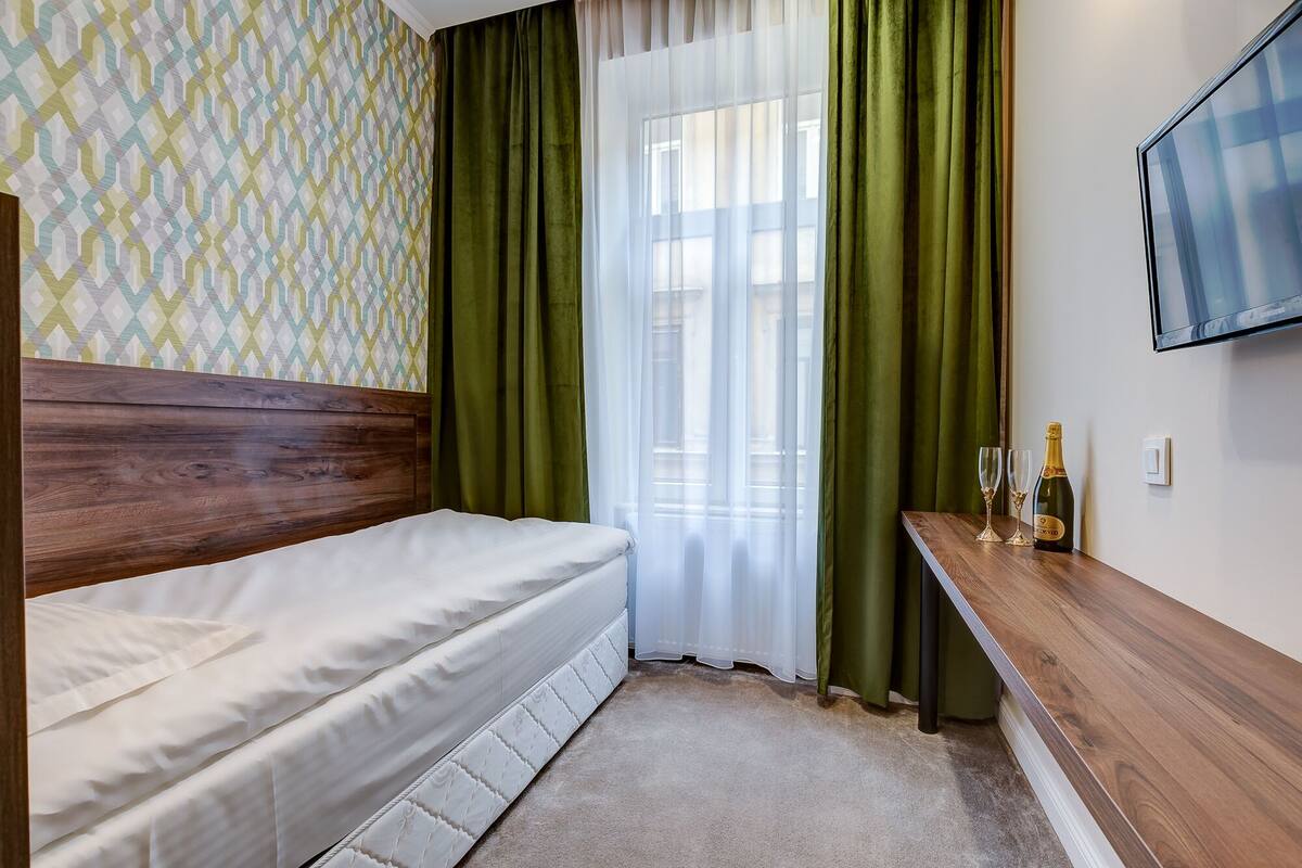 A bedroom with a wooden headboard, a bed with white bedding, green curtains, a small table with glasses and a bottle, and a TV mounted on the wall.