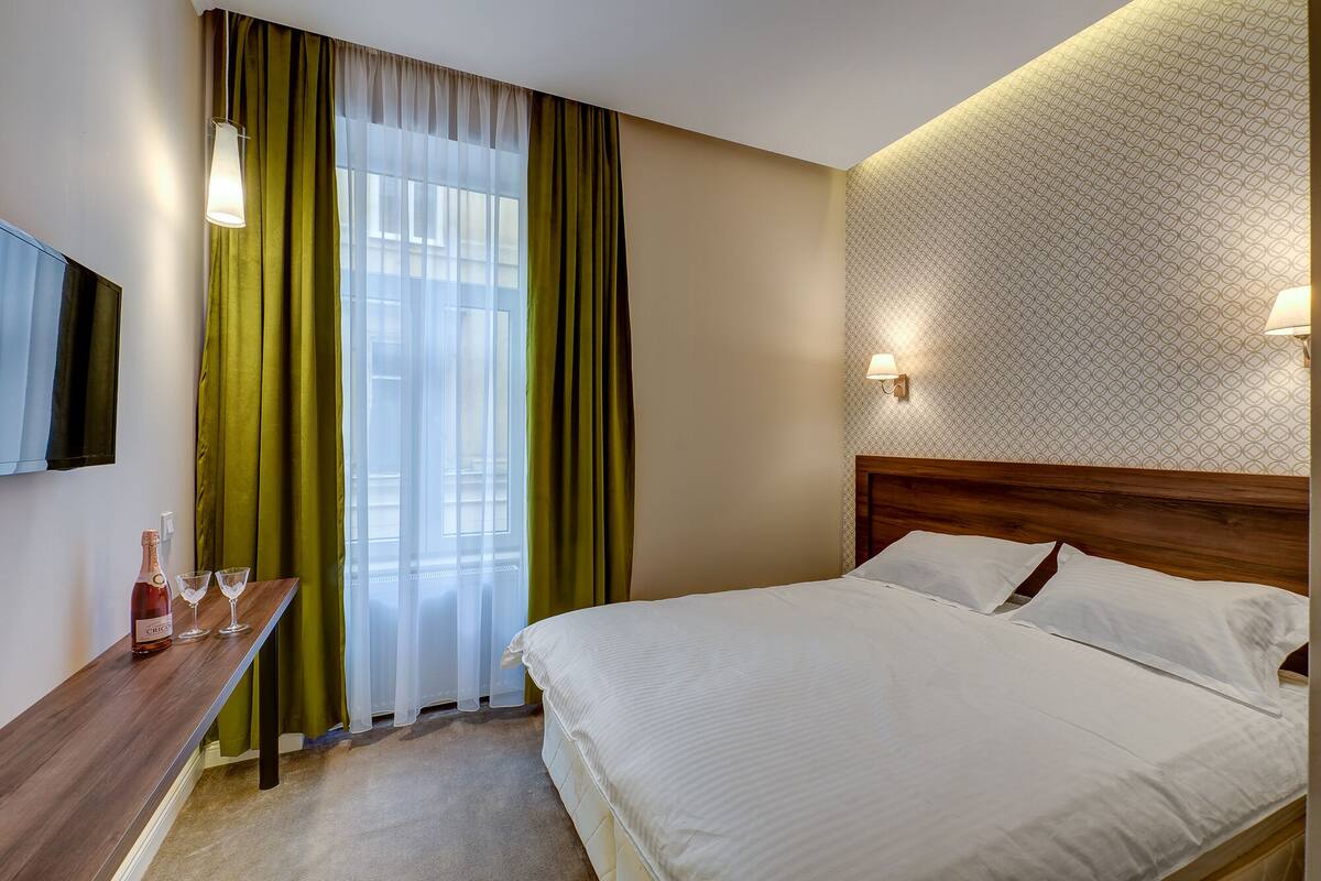 A hotel room with a bed, a wooden headboard, a bedside table with a bottle and glasses, a TV mounted on the wall, and a window with green curtains.