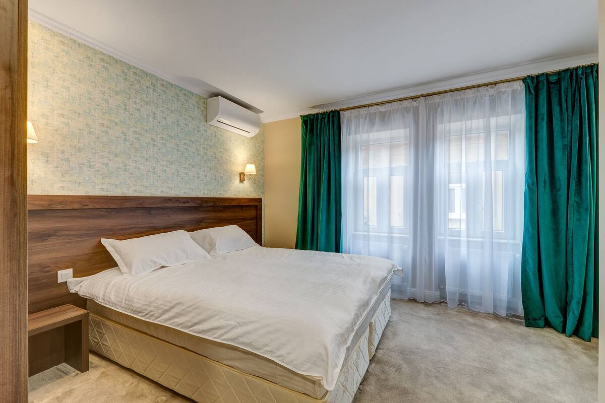 A neatly made bed with white linens in a room with a wooden headboard, a wall-mounted air conditioner, and a window with teal curtains.