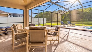 Un patio couvert avec une table et des chaises, un ventilateur de plafond, et une vue sur une piscine et la verdure environnante.