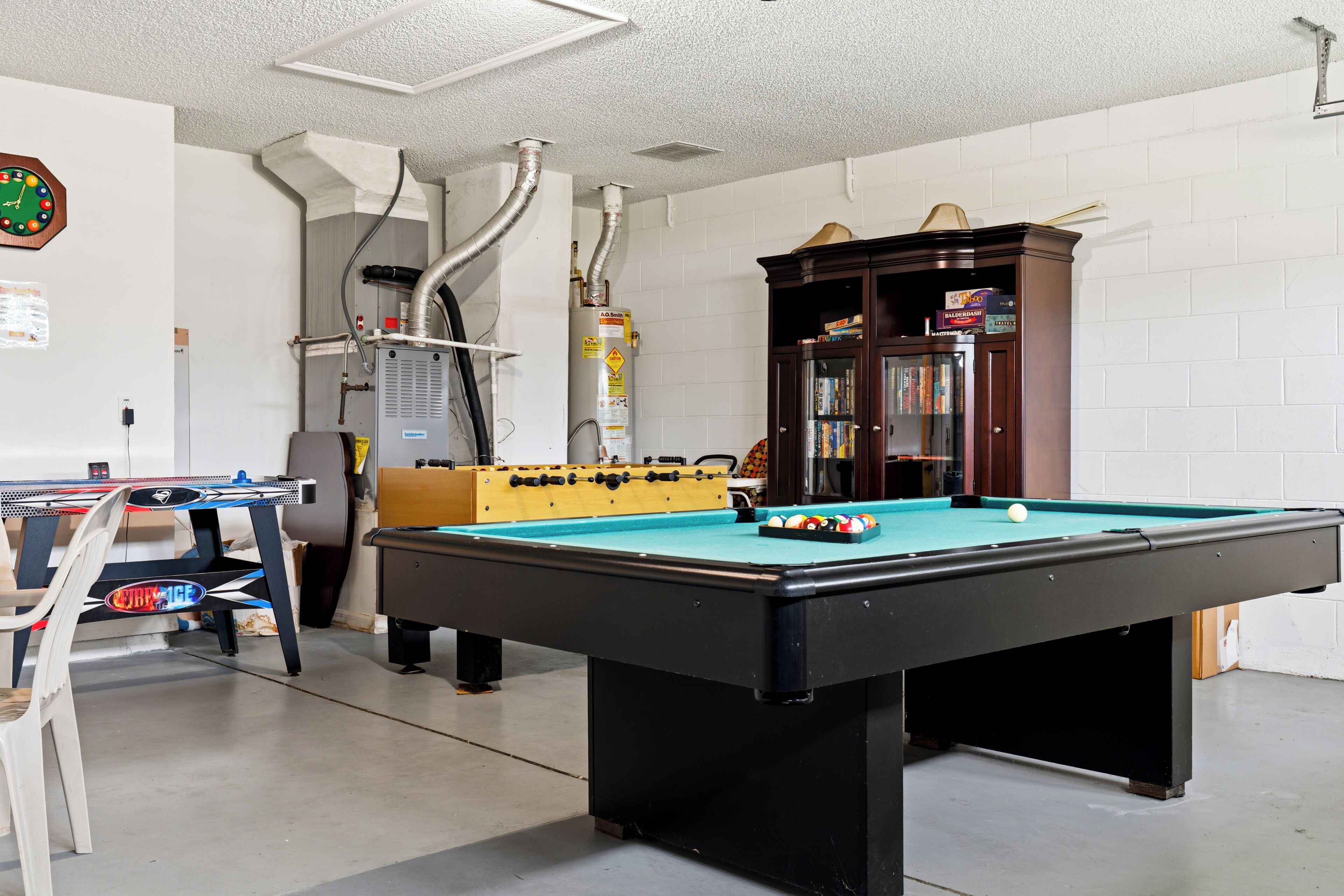 A pool table with a foosball table and a bookshelf in a room with white walls and a ceiling vent.