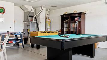 A pool table with a foosball table and a bookshelf in a room with white walls and a ceiling vent.