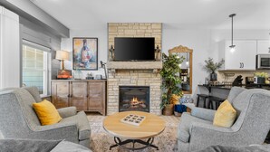 A living room with grey sofas, a wooden coffee table, a fireplace, a flat-screen TV, and a kitchen area in the background. - Lakefront Luxury:
NoStairs/King beds
Wheelchair Accessible
Families/Couples/Pets (Branson)