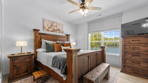 A bedroom with a wooden bed, nightstands, a bench, a ceiling fan, and a large window with a view of greenery. - Lakefront Luxury:
NoStairs/King beds
Wheelchair Accessible
Families/Couples/Pets (Branson)