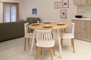 A dining area with a round table, white chairs, and a kitchen in the background. - Coastal Getaway: Roomy Beachside Apartment! (Nea Makri)