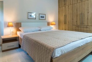 A bedroom with a bed, two bedside lamps, a wooden wardrobe, and two framed pictures on the wall. - Coastal Getaway: Roomy Beachside Apartment! (Nea Makri)