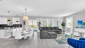 A modern living room with a grey sectional sofa, a dining area with white chairs, and a kitchen with white cabinets and a black countertop.