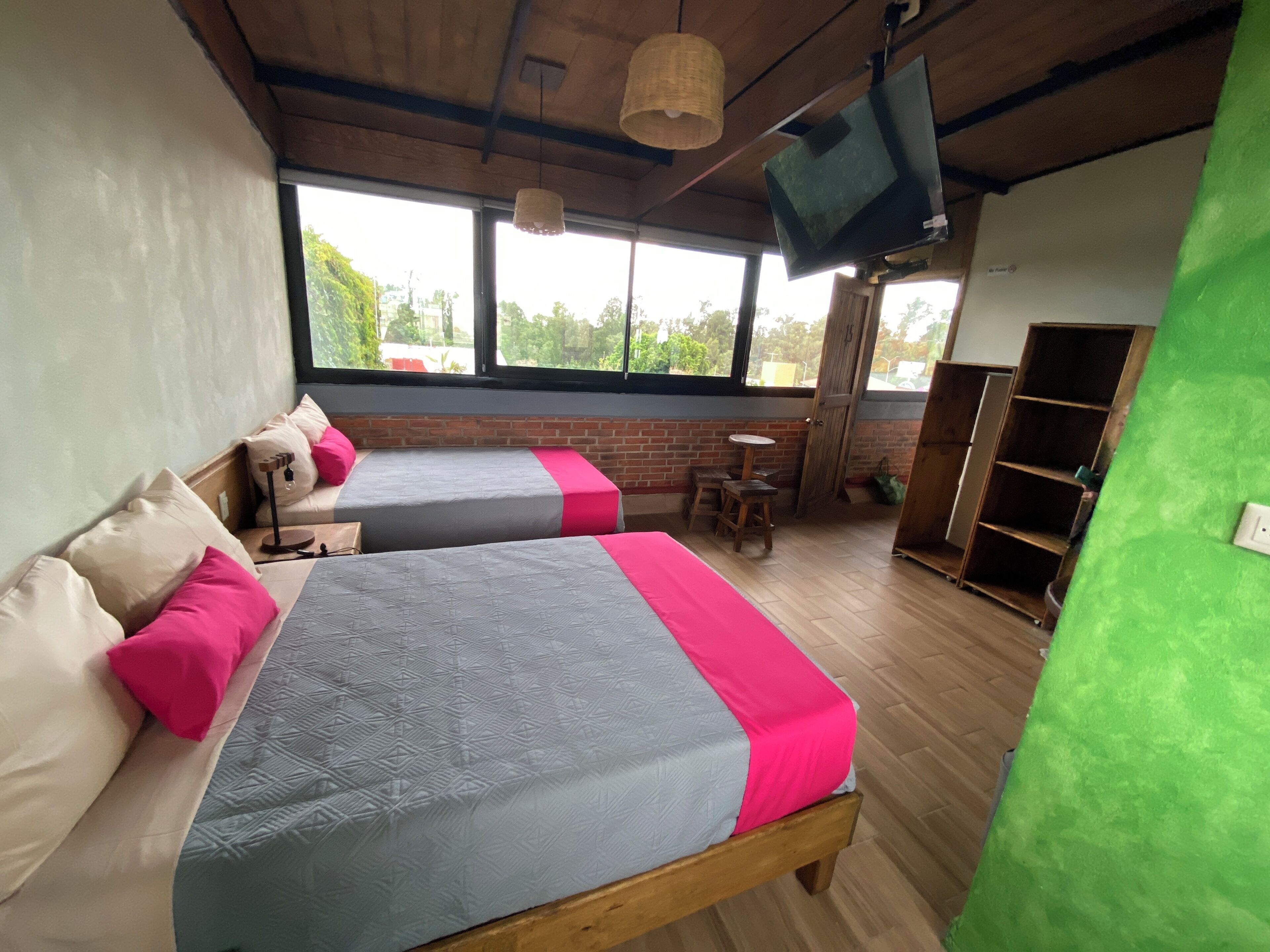 A room with two beds, a small table, a television, and a view of the outdoors through large windows.