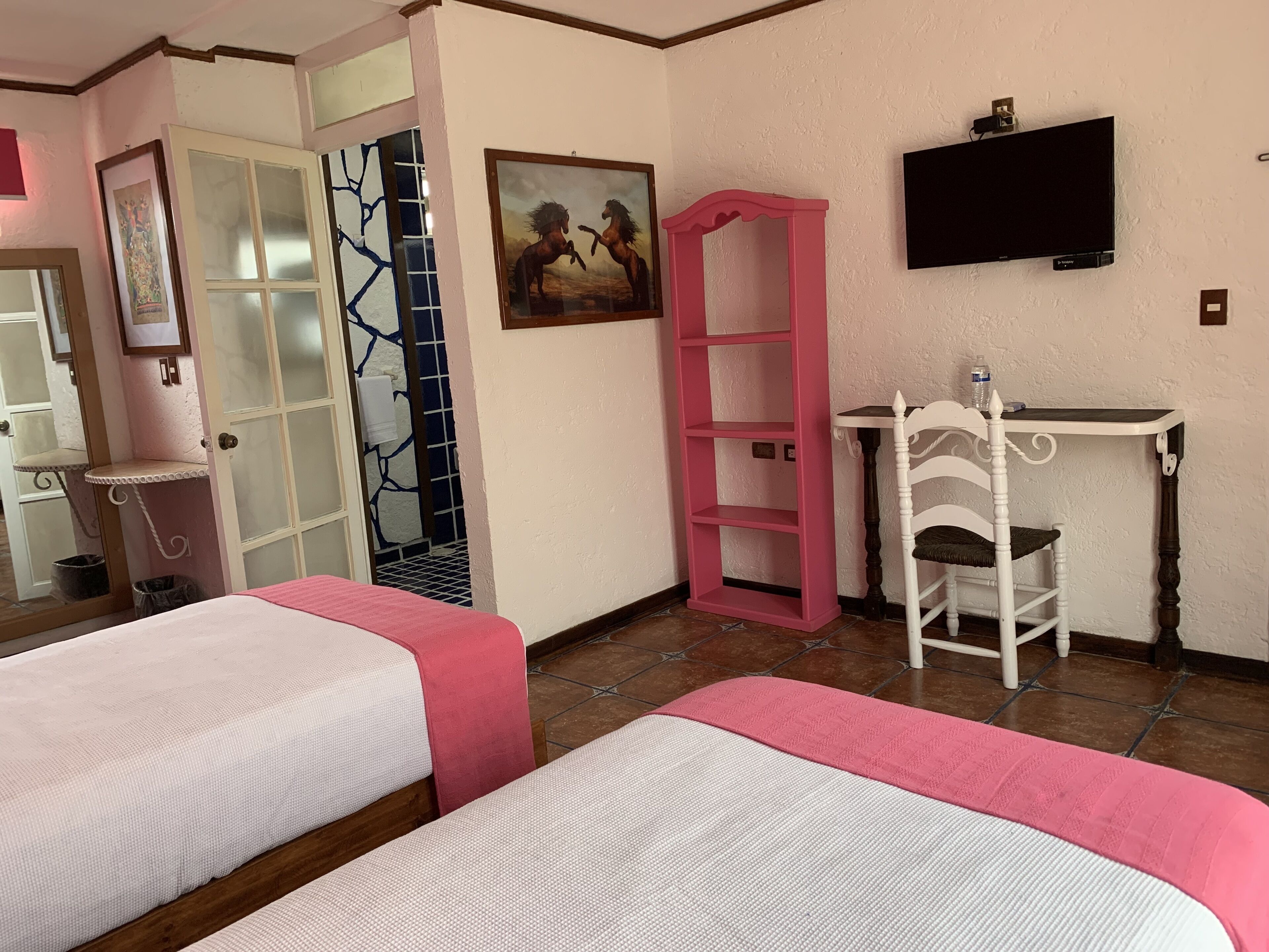 A hotel room with two beds, a pink chair, a desk, a TV, and a painting.