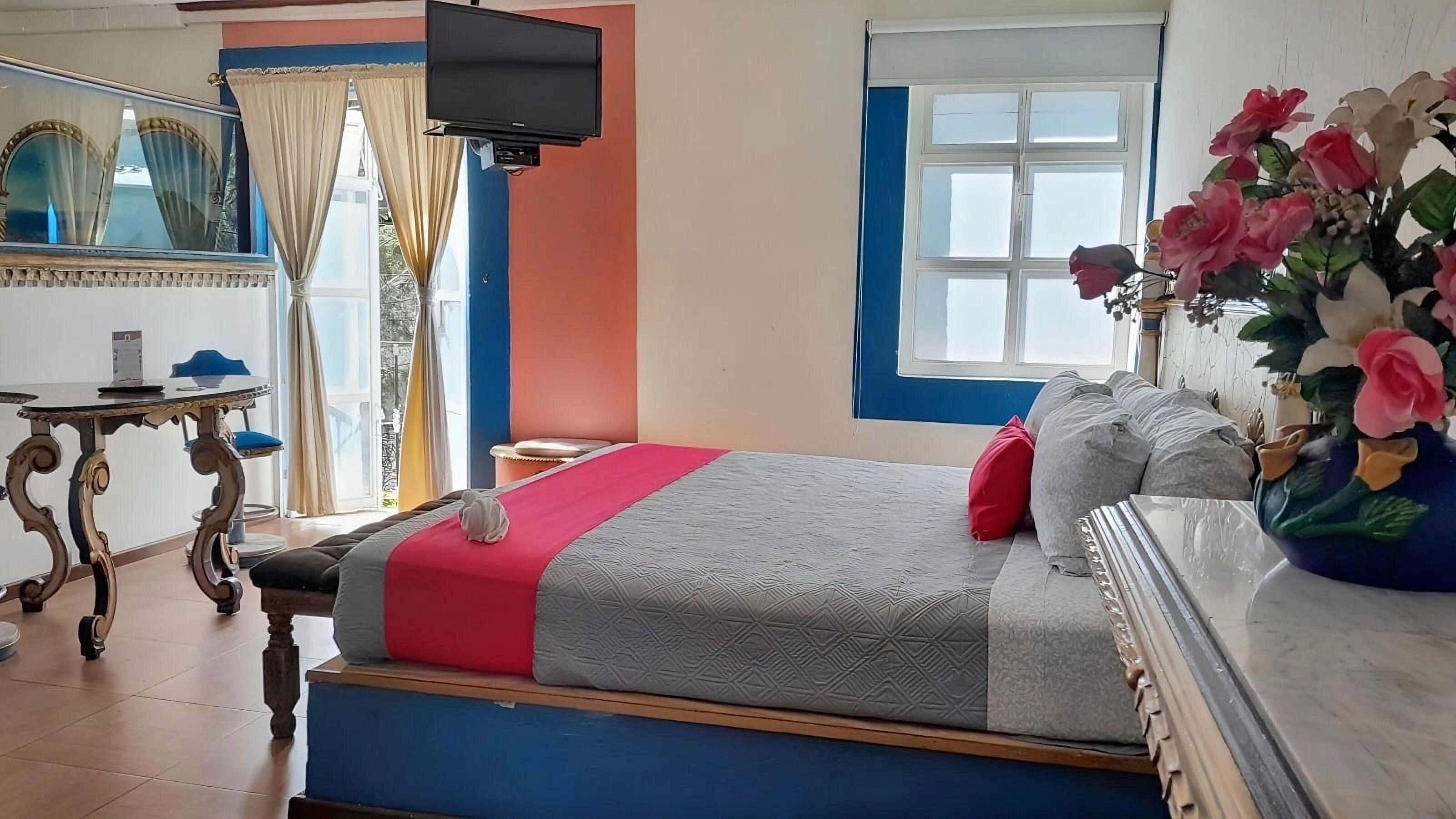 A bedroom with a bed, a TV, a window with curtains, a dresser, and a vase with flowers.