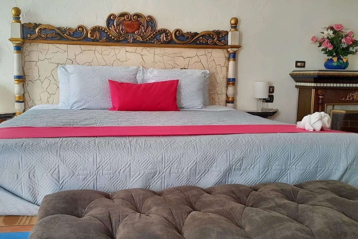 A bed with a patterned bedspread, a red pillow, and a tufted ottoman in the foreground.
