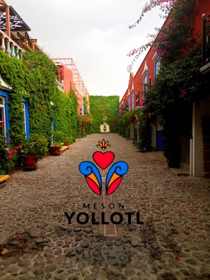 A cobblestone street with buildings on either side, a logo with hearts and the text "MESON YOLOTL" in the center, and a stone fountain in the distance.