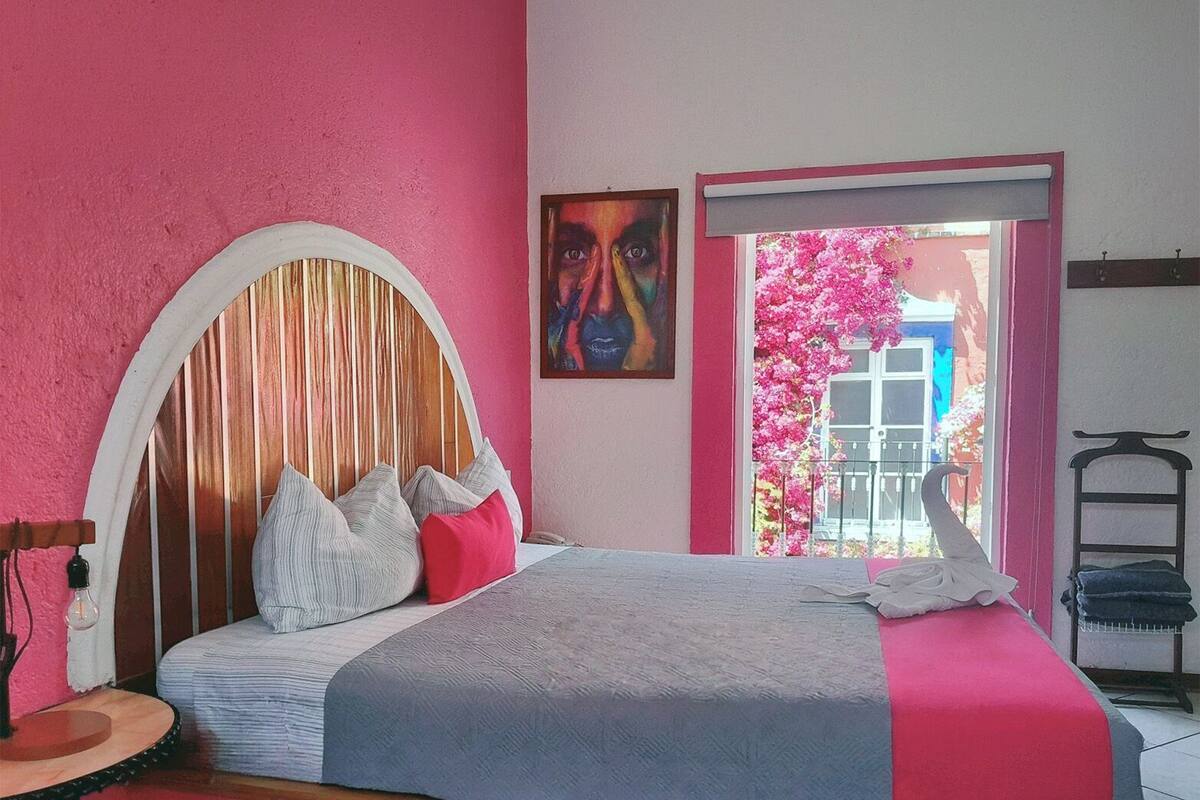 A bedroom with a pink wall, a wooden headboard, a bed with pillows, a painting, and a view of pink flowers through a door.