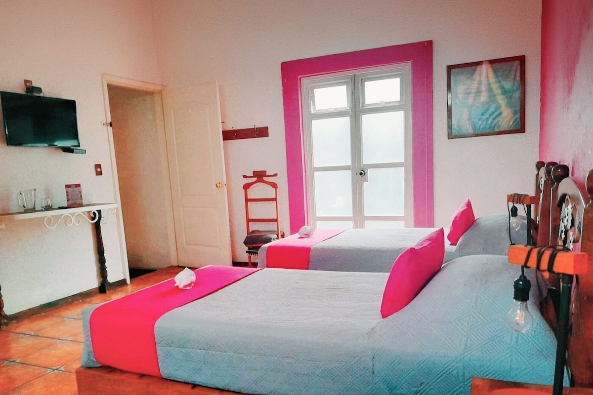 A room with two beds, a TV, a window with pink trim, and a painting on the wall.