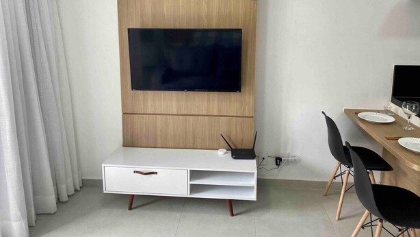 A modern hotel room with a flat-screen TV mounted on a wooden panel wall, a white console table with a drawer, a dining area with a wooden table and black chairs, and a white curtain on the left.