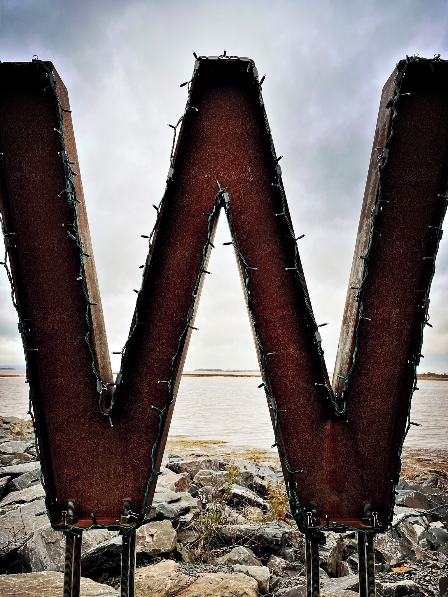 A large metal structure resembling the letter 'W' with a rugged, industrial design, set against a backdrop of a rocky shoreline and a body of water.