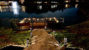 Une vue nocturne d’un chemin pavé menant à une zone fermée par une grille, où se trouvent une fontaine et une étendue d’eau reflétant les lumières.