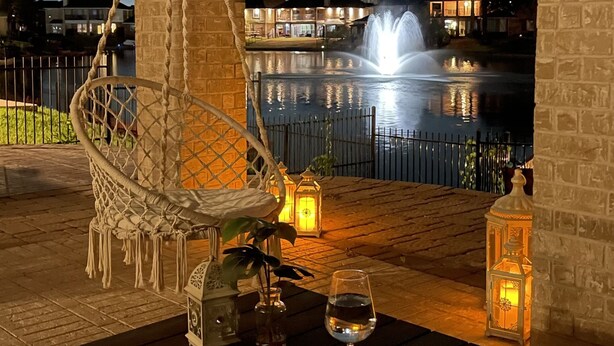 Une table en bois, un verre d’eau, une lanterne et un fauteuil suspendu sur une terrasse aux murs de briques, avec vue sur une fontaine et des bâtiments en arrière-plan.