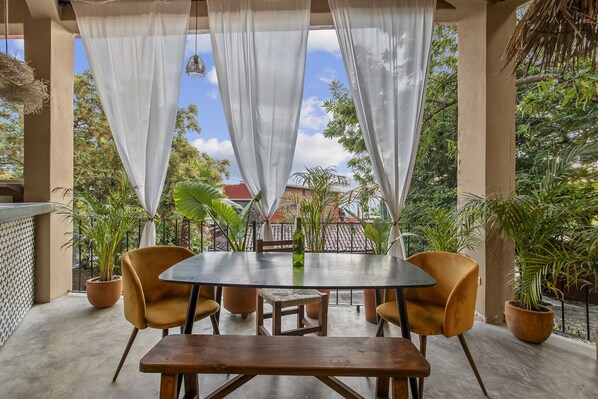 A covered outdoor dining area with a round table and four chairs, surrounded by potted plants and a view of buildings in the background. - Mountain Paradise Retreat: Casanevo-Tepoztlan (Tepoztlán)