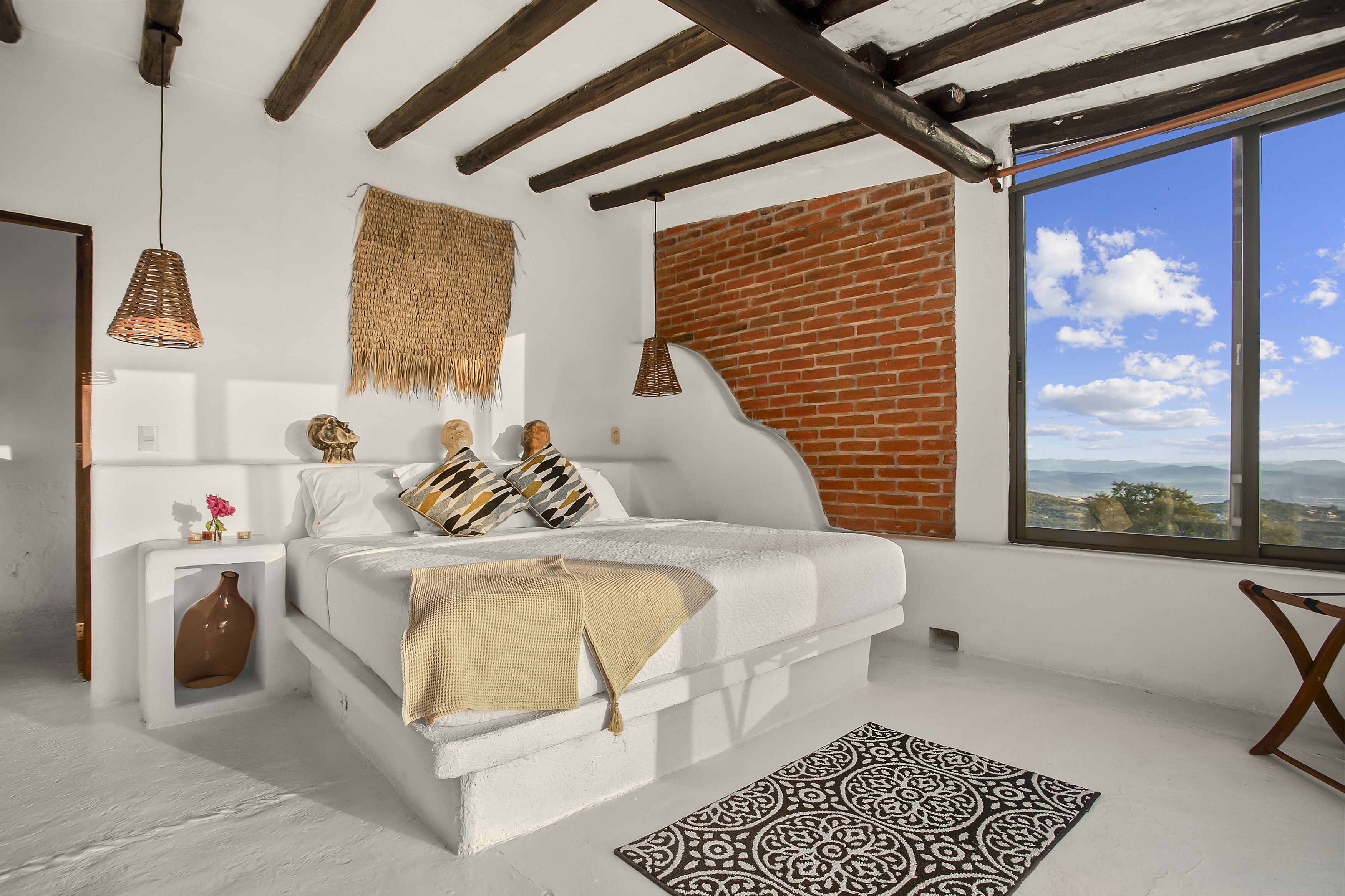 Un dormitorio moderno con una cama blanca, una pared de ladrillo y un amplio ventanal con vistas al cielo y al paisaje.
