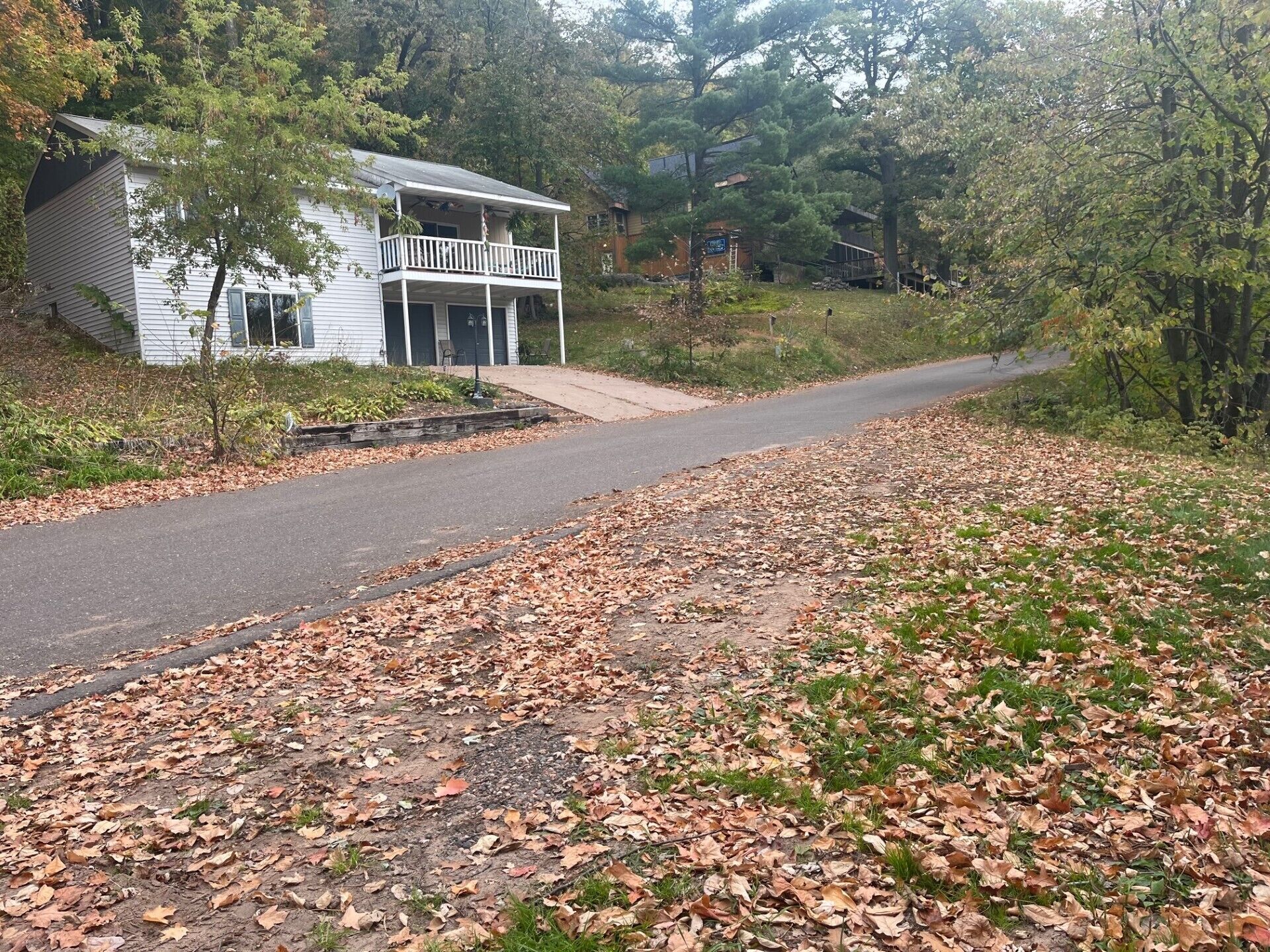 Una casa de dos plantas con balcón, rodeada de árboles y un camino de entrada cubierto de hojas caídas.