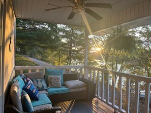 Une véranda couverte avec un ventilateur de plafond, un canapé en osier avec des coussins et une vue sur les arbres et une route.
