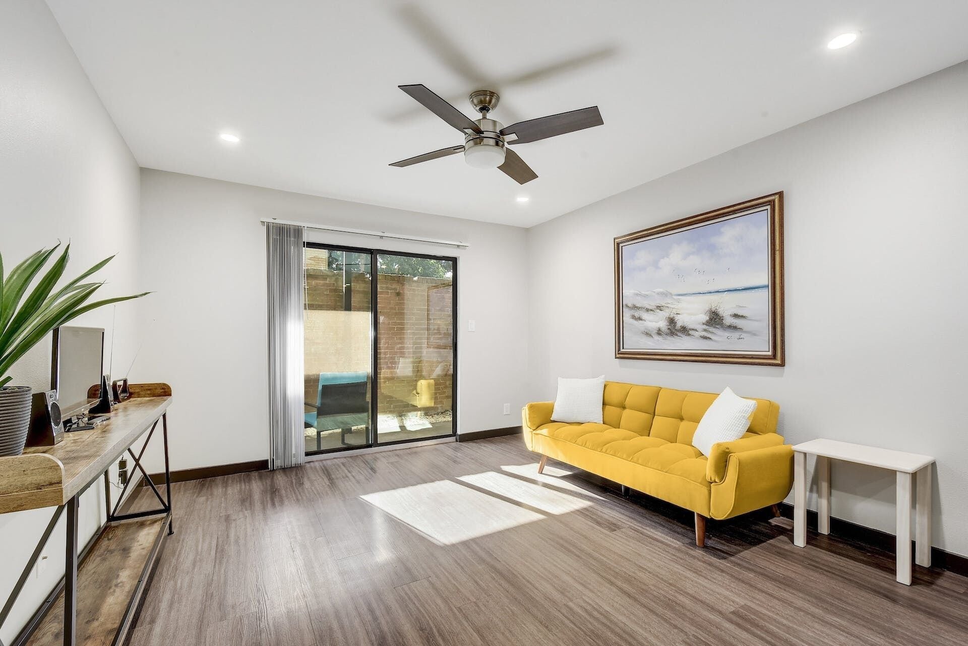 Uma sala de estar moderna com um sofá amarelo, um ventilador de teto, uma janela grande e um quadro na parede.