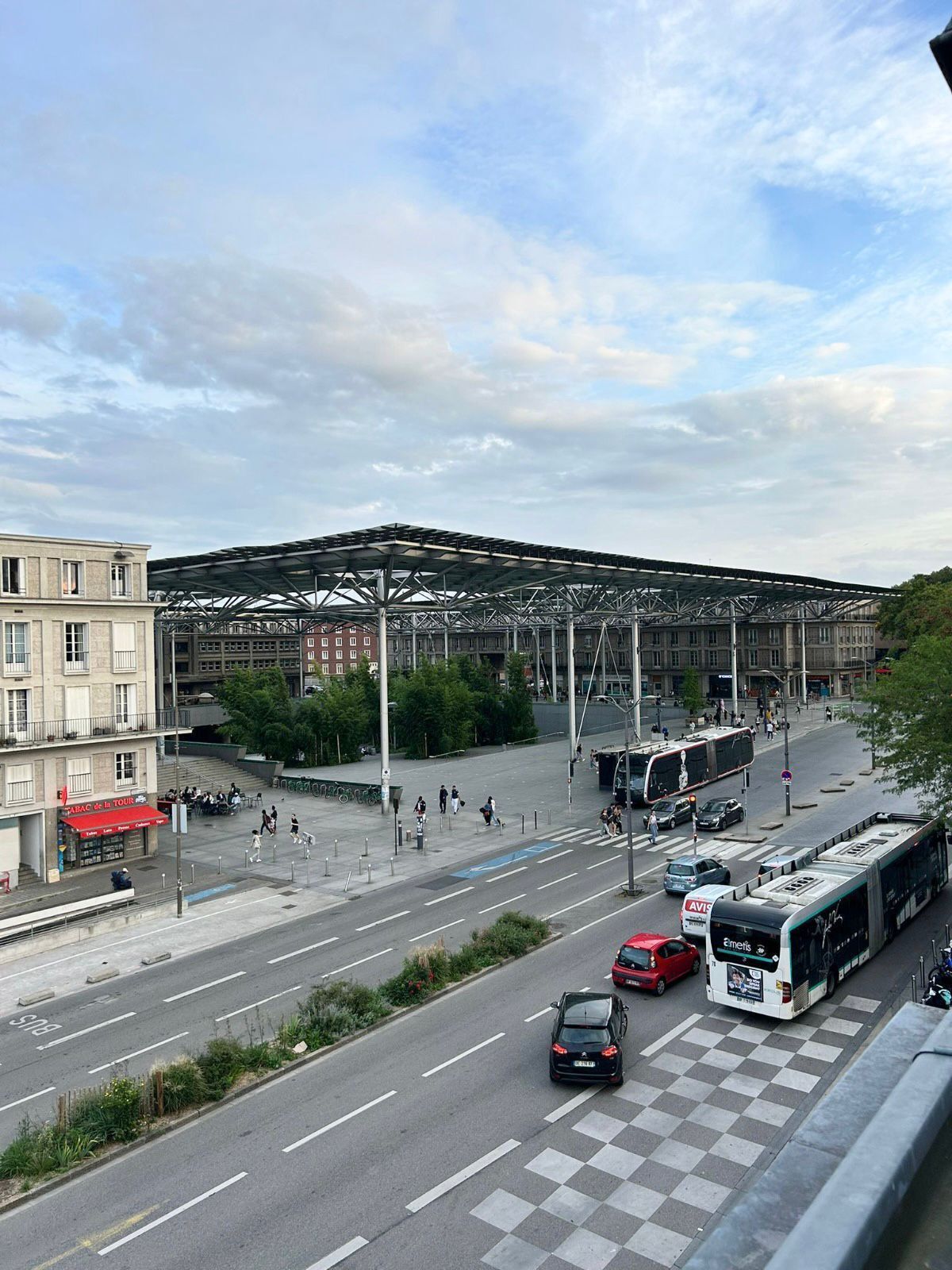 A street view with a bus stop, a pedestrian crossing, and a large building with a metal framework.