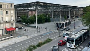 Une vue de rue avec un arrêt de bus, un passage piéton et un grand bâtiment doté d’une structure métallique.