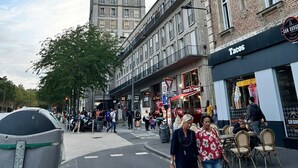 Une rue bordée d’un grand immeuble, d’un restaurant appelé « Tacos » et de piétons.
