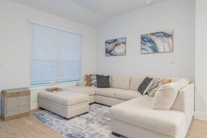 A modern living room with a sectional sofa, a small ottoman, and a blue patterned rug. There are two abstract paintings on the wall and a wicker basket on the floor.