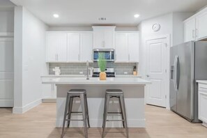 A modern kitchen with white cabinets, a central island with stools, stainless steel appliances, and a clock on the wall.