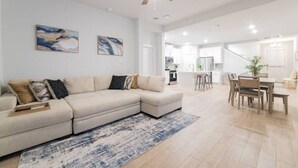 En rummelig stue med en stor hjørnesofa, et spiseområde med et træbord og stole, og et køkken i baggrunden.