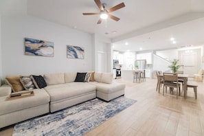 A spacious living room with a large sectional sofa, a dining area with a wooden table and chairs, and a kitchen in the background.